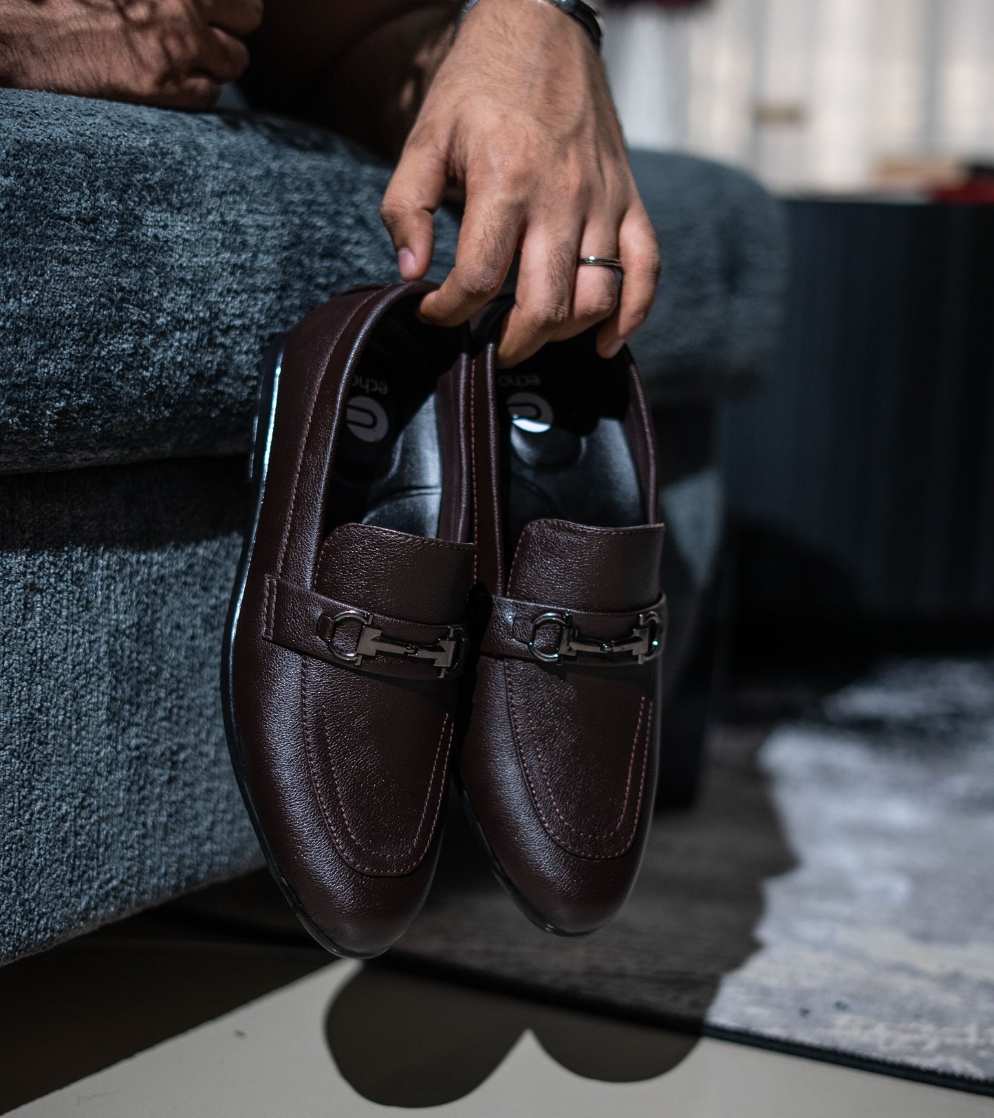 Brown leather loafers held by a person sitting on a gray couch.