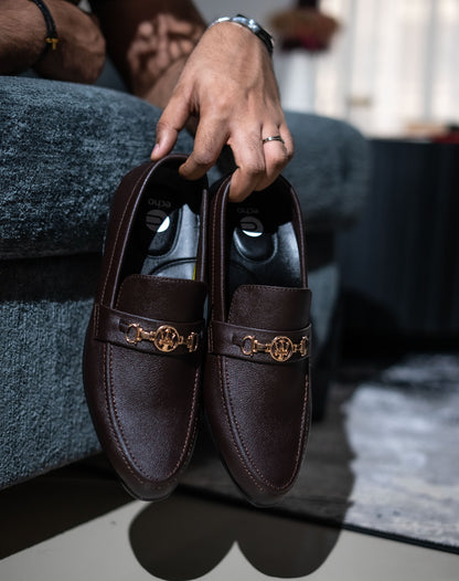 Brown loafers with gold accents held by a hand on a blurred background