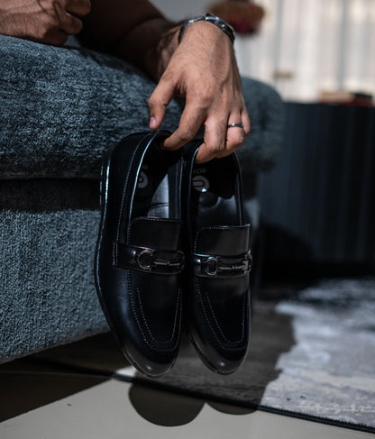 Person holding a pair of black loafers on a blurred indoor background