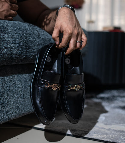 Person holding a pair of black loafers with gold accents on a checkered floor.