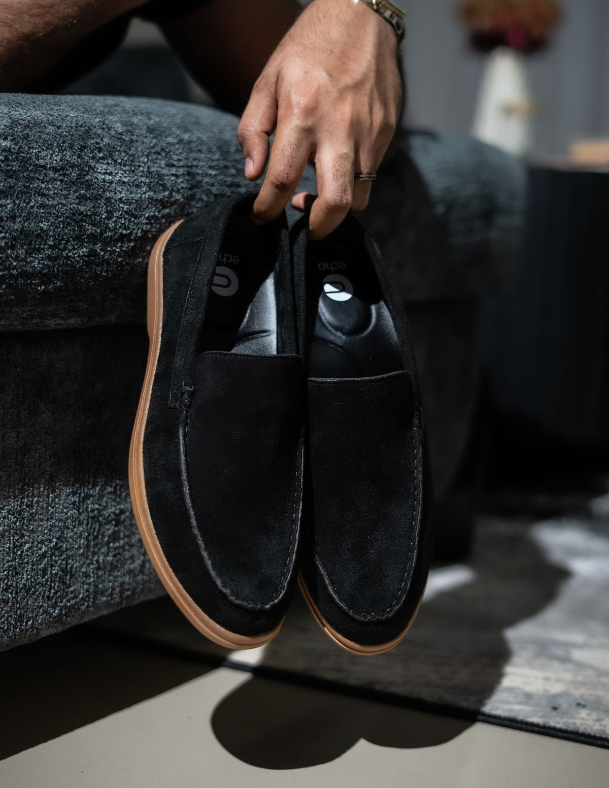 Person holding a pair of black loafers with brown soles on a gray couch.