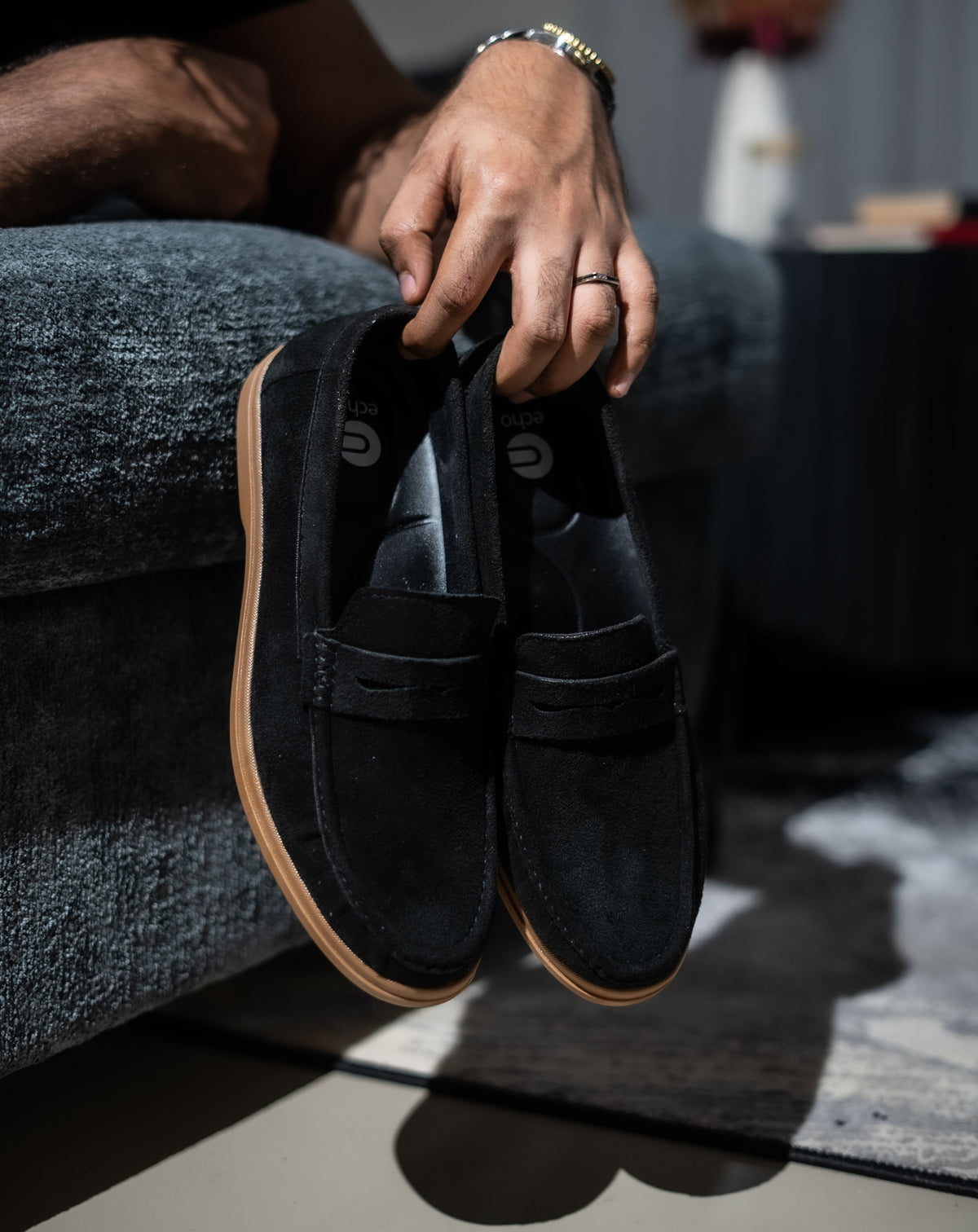 Person holding a pair of black loafers with brown soles on a gray couch.