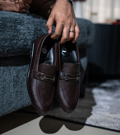 Brown leather loafers held by a person sitting on a gray couch.