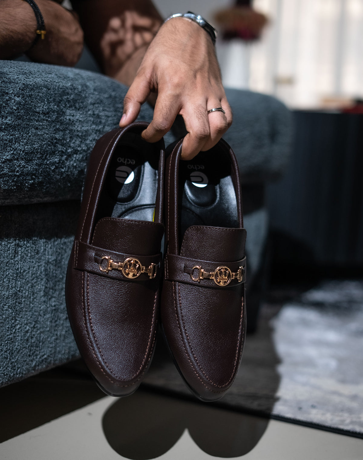 Brown loafers with gold accents held by a hand on a blurred background