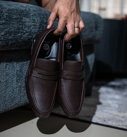 Brown leather loafers held by a person sitting on a couch.