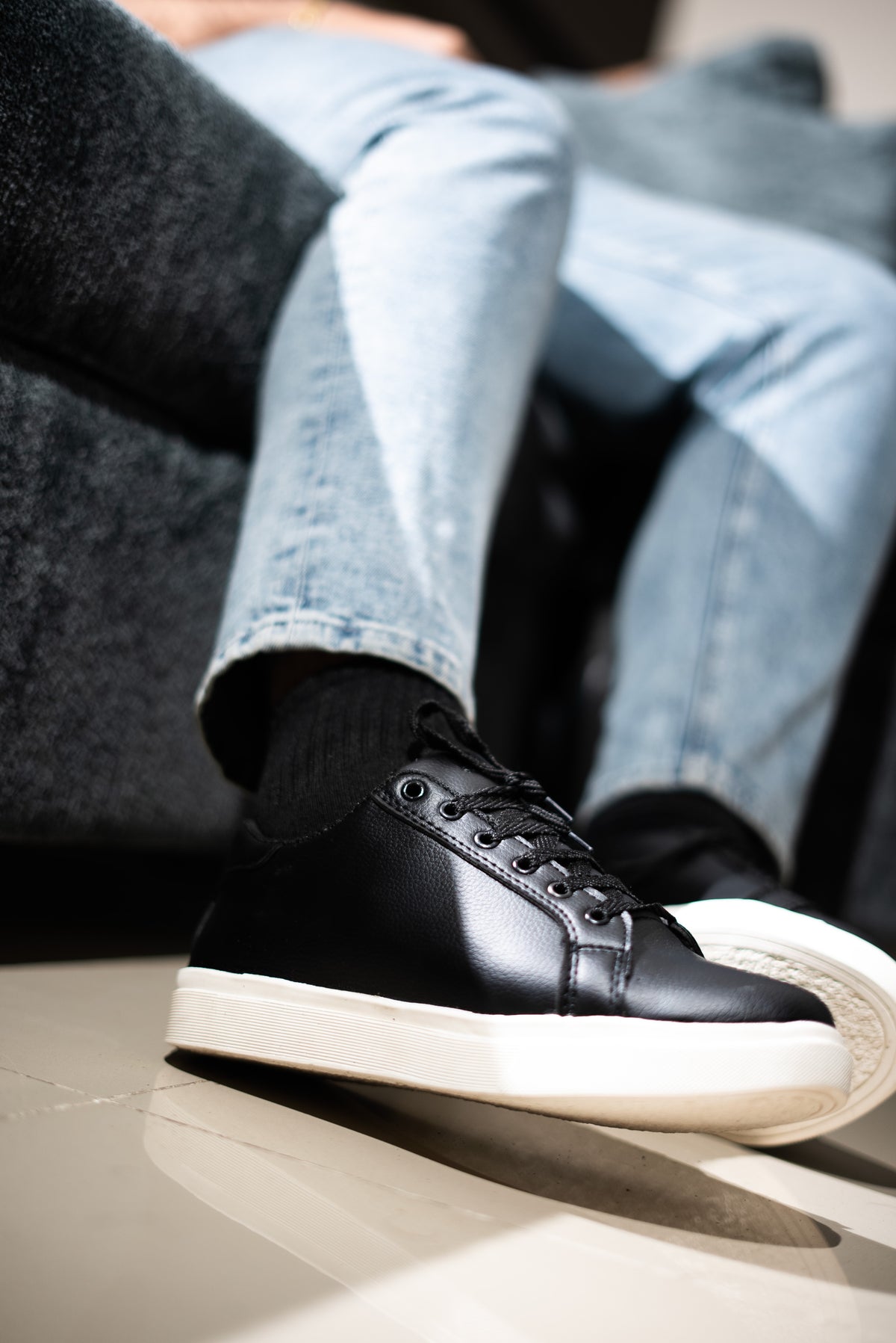 Person wearing black sneakers with white soles and light blue jeans sitting on a gray couch.